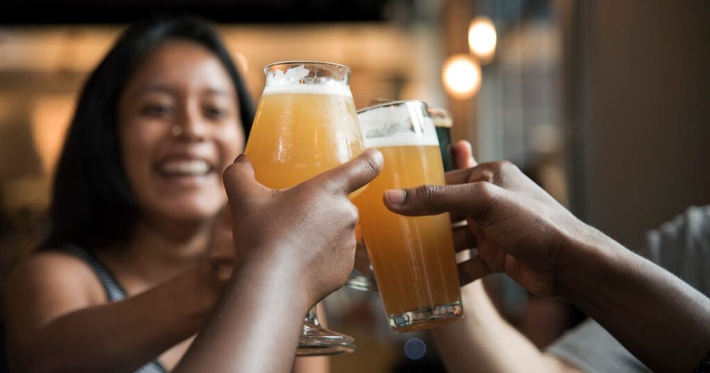 Glass of golden non-alcoholic beer with perfect white head on wooden table, hop cones and barley grains